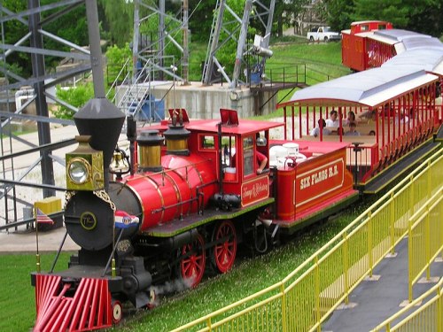 A red steam train with a black smokestack, pulling a passenger car at an amusement park, surrounded by green grass.