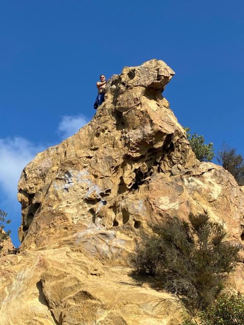 A person stands triumphantly on top of a large rock formation against a clear blue sky.