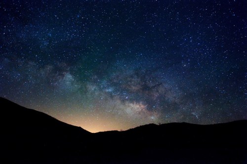 A starry night sky filled with stars and the Milky Way, silhouetted against dark mountains and a faint horizon glow.