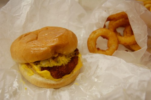 A sandwich with a breaded patty, cheese, and pickles, next to a serving of onion rings, all wrapped in paper.