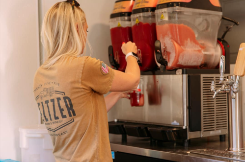 A woman in a tan shirt serves a slushy drink from a machine in a casual setting.