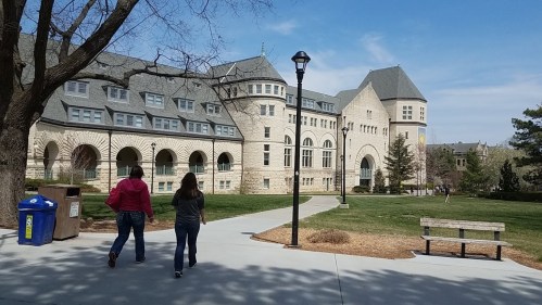 Two people walk along a path in front of a large stone building on a sunny day, with trees and a bench nearby.