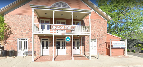 A brick building with a sign reading "Opera Hall" and a porch, surrounded by trees and a sidewalk.