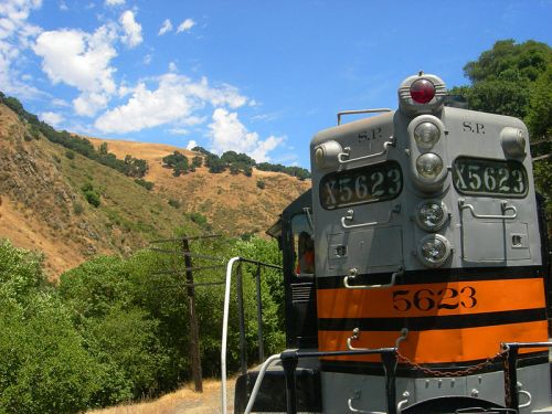 A vintage train engine with a black and orange design, set against a backdrop of green hills and blue sky.