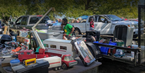 A busy scene at a yard sale with various items for sale, including toys, tools, and vehicles, with people browsing.