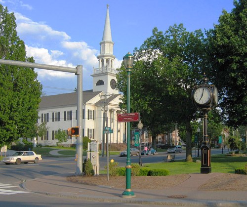 A church with a tall steeple stands near a clock tower in a green park-like area, with trees and a clear blue sky.