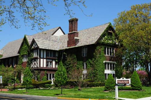 A large, ivy-covered Tudor-style house with a steep roof and manicured gardens on a sunny day.