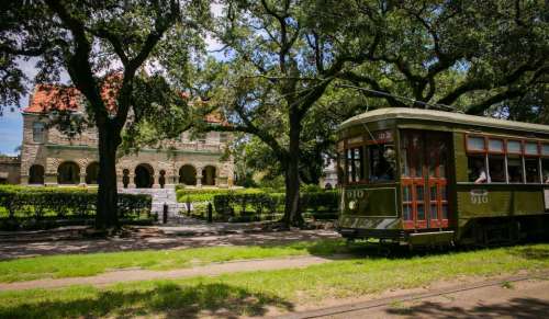 A vintage green streetcar parked beside a historic mansion, surrounded by lush trees and greenery.