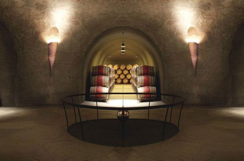 A dimly lit wine cellar featuring stacked barrels and a circular table in the foreground. Warm, rustic ambiance.