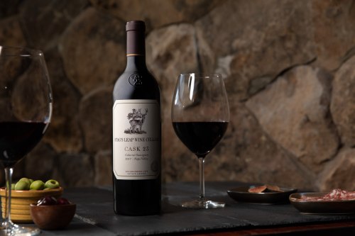 A bottle of red wine with two glasses, olives, and charcuterie on a stone table.