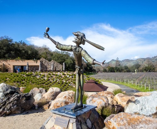 A bronze statue of a joyful figure holding a glass, set against a vineyard and blue sky backdrop.