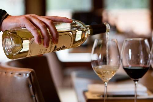 A hand pours white wine from a bottle into a glass, with another glass of red wine in the background.