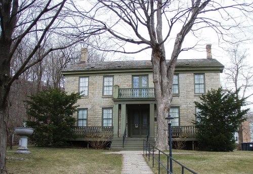 Historic stone house with green trim, surrounded by trees and a lawn, featuring a front porch and balcony.