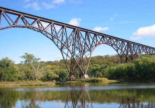 A large metal bridge spans over a calm river, surrounded by lush greenery and a clear blue sky.