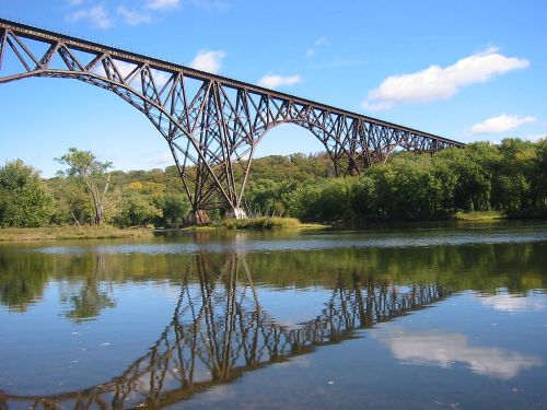 A large metal bridge spans over a calm river, reflecting its structure in the water, surrounded by trees and blue sky.