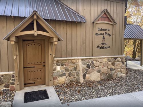 A wooden building with a stone foundation, featuring a welcoming sign for "Stoney Ridge Lighthouse" and a decorative door.