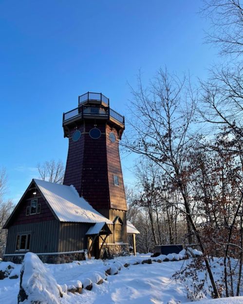 A tall, wooden lighthouse surrounded by snow, with a clear blue sky and trees in the background.