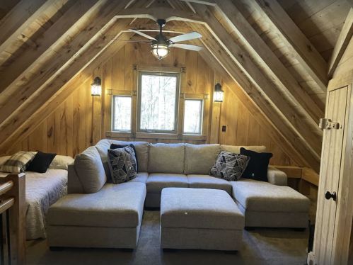 Cozy cabin interior with a large sectional sofa, wooden walls, and a ceiling fan, featuring natural light from windows.