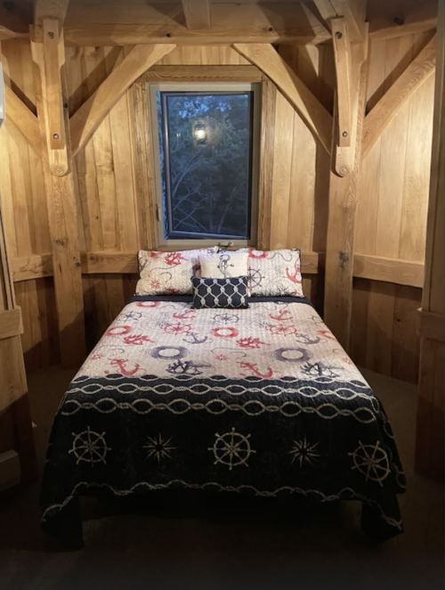 A cozy bedroom with a wooden interior, featuring a bed covered in a nautical-themed quilt and a window with a view.