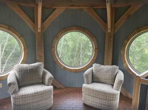 Cozy interior of a circular room with two plaid armchairs and large round windows overlooking greenery.