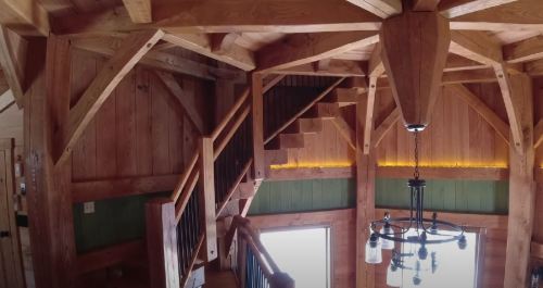 Interior view of a wooden cabin featuring exposed beams, a staircase, and a chandelier. Warm, rustic ambiance.
