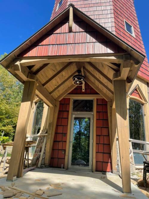 A wooden structure with a red shingle roof, featuring a large entrance and exposed beams, surrounded by trees.