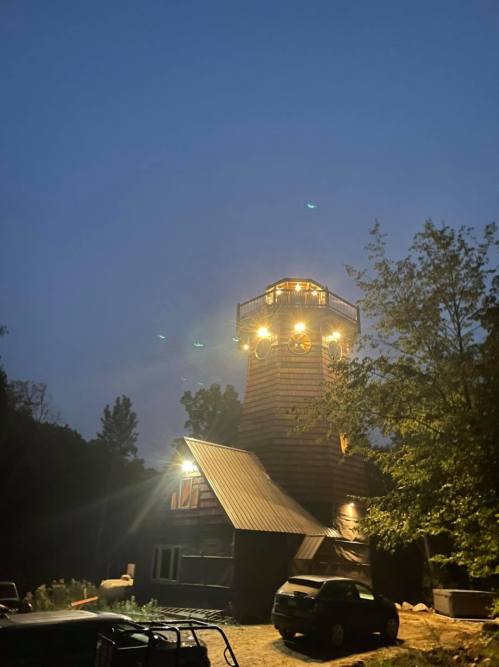 A tall, illuminated tower stands against a twilight sky, surrounded by trees and parked vehicles.
