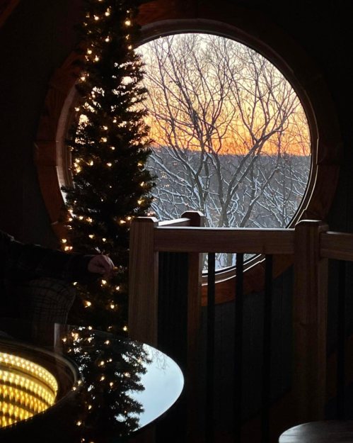 A cozy interior with a Christmas tree, a round window showing a sunset over snowy trees outside.