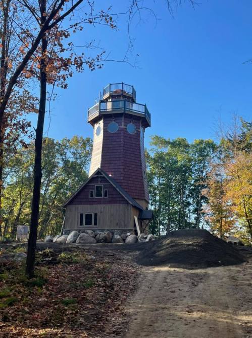 A tall, unique lighthouse-style building surrounded by trees and a dirt path, with a pile of dirt in the foreground.