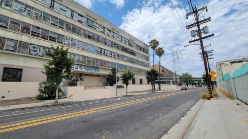 Abandoned building with graffiti, palm trees, and a quiet street under a cloudy sky. Power lines in the background.