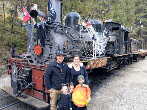 A family poses in front of a decorated steam train for Halloween, with kids in costumes and festive decorations.