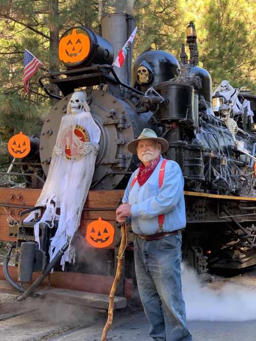 A man in a hat and suspenders stands beside a decorated steam train with Halloween decorations and skeletons.