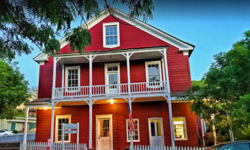 A red two-story house with a white porch and picket fence, surrounded by greenery at dusk.