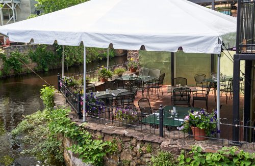 Outdoor dining area under a white tent by a river, featuring tables, chairs, and colorful flower pots.