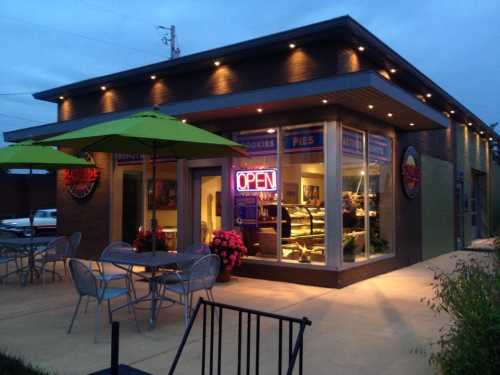 A cozy bakery with outdoor seating, illuminated "OPEN" sign, and colorful umbrellas at dusk.