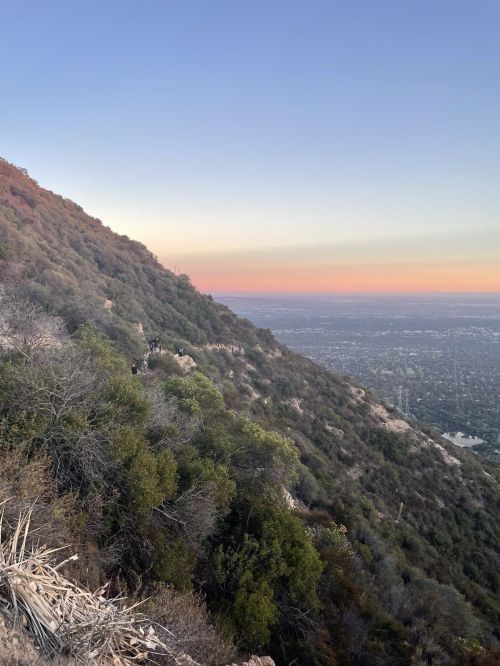 A scenic view of a mountainside at sunset, overlooking a city with a gradient sky in soft hues.