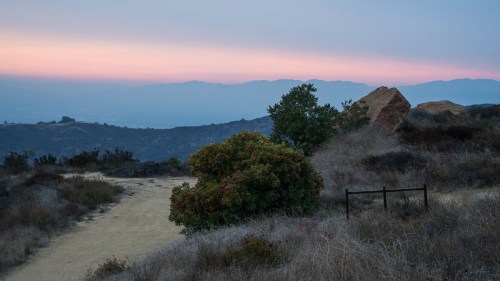 A serene landscape at dusk, featuring a dirt path, shrubs, and distant mountains under a colorful sky.