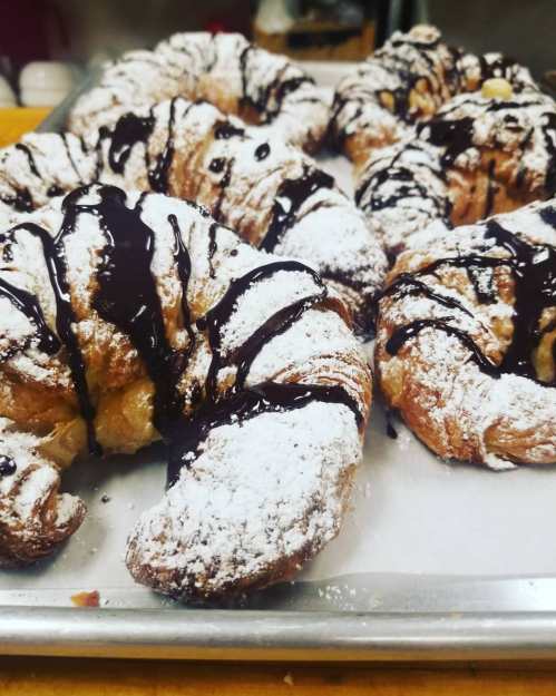 Freshly baked croissants dusted with powdered sugar and drizzled with chocolate sauce on a baking tray.
