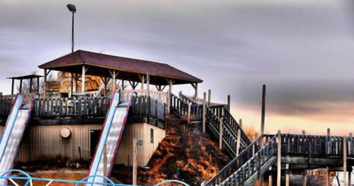 A wooden play structure with slides on a hill, set against a cloudy sky at dusk.