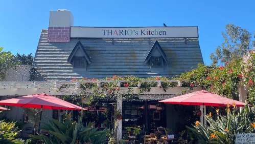 A charming restaurant exterior with a sign reading "THARIO's Kitchen," surrounded by greenery and red umbrellas.