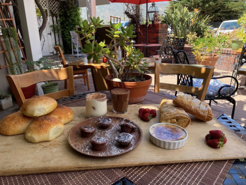 A wooden table outdoors displays various baked goods, including rolls, pastries, and desserts, surrounded by plants.