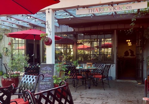 Outdoor dining area of Thario's Kitchen, featuring tables, red umbrellas, and a chalkboard menu. Cozy and inviting atmosphere.
