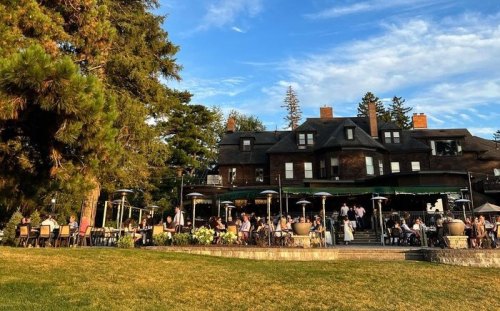 A large historic building with outdoor dining, surrounded by trees and a grassy area under a blue sky.