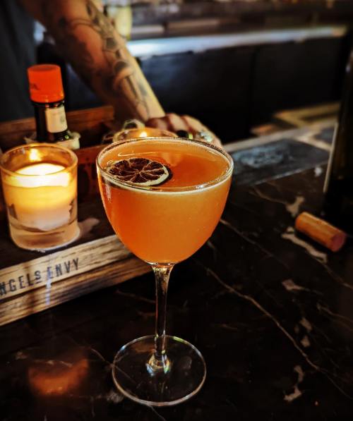 A cocktail in a stemmed glass, garnished with a dried citrus slice, sits on a dark marble bar with a candle in the background.