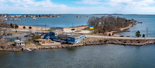 A coastal view featuring a blue building, sandy beach, and calm waters under a clear sky.