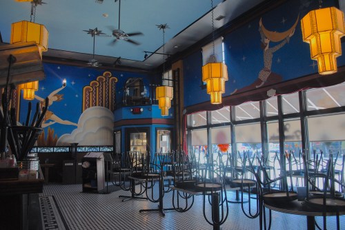 A colorful, empty café interior with artistic murals, ceiling fans, and stacked chairs on tables.