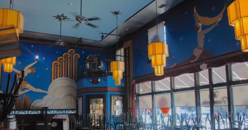 Interior of a colorful restaurant featuring blue walls, artistic murals, and vintage light fixtures. Chairs are stacked.