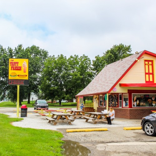 A colorful roadside eatery with picnic tables outside, surrounded by greenery and a sign that reads "The Thirsty Farmer."
