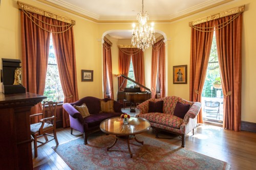 Elegant living room with a grand piano, plush seating, and large windows draped in rich curtains, illuminated by a chandelier.