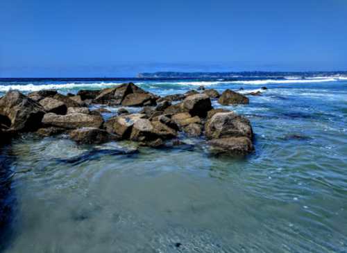 Rocky shoreline with clear blue water and gentle waves under a bright blue sky.